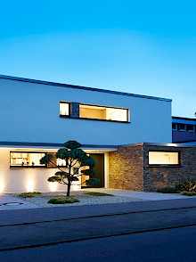 Moderner Vorgarten in Paderborn mit Kiesfläche, formgeschnittener Kiefer und dezent beleuchteter Hausfassade.