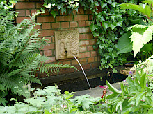 Brunnen an einer Klinkermauer mit mit umrahmender Bepflanzung aus schattenverträglichen Stauden.