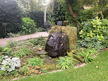 Großer Natursteinfindling als Quellstein mit Schattenstauden und Farnen in einem Hausgarten in Paderborn.