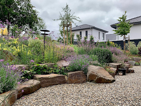 Hanggestaltung in einem Garten in Lichtenau aus Grauwackefindlingen und intensiver Staudenbepflanzung. Angrenzend daran eine provisorische Terrasse aus Lippkies.