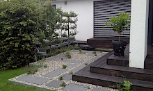 Garten in Paderborn mit einer Stufenanlage und angrezender Terrasse aus geöltem Holz sowie einem bepflanzten Kiesbereich mit Schirttplatten aus Basaltlava.