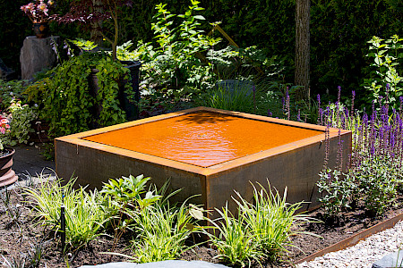 Eckiges Wasserspiel aus Cortenstahl in einem Staudenbeet eines Privatgartens in Paderborn.
