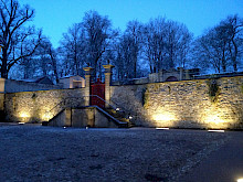 Historische Mauer mit einem roten Tor im Kloster Dalheim in Lichtenau, die von länglichen Bodenstrahlern angeleuchtet werden.