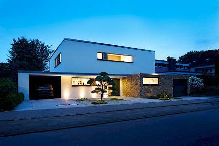 Puristisch gestalteter Vorgarten an einem modernen Einfamilienhaus in Paderborn mit beleuchteter Formkiefer und stimmungsvoller Fassadenbeleuchtung in der Abenddämmerung.