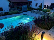 Abendstimmung in eienm Privatgarten mit beleuchtetem Pool und Pollerleuchte an einer Treppe.