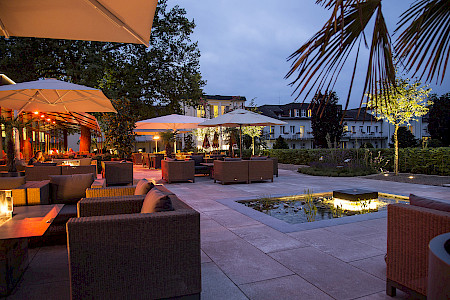 Gastronomieterrasse in Bad Lippspringe im Abendlicht mit unterschiedlichen Beleuchtungselementen. Im Fokus steht der beleuchtete Quellestein in der Mitte des Wasserbeckens.